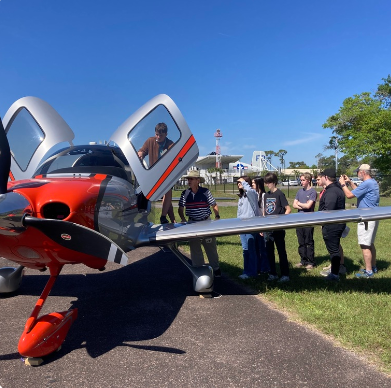 Group of students and adults gathered around a small red and silver airplane with open doors on a sunny day. One person is seated inside the cockpit while others observe and take photos.