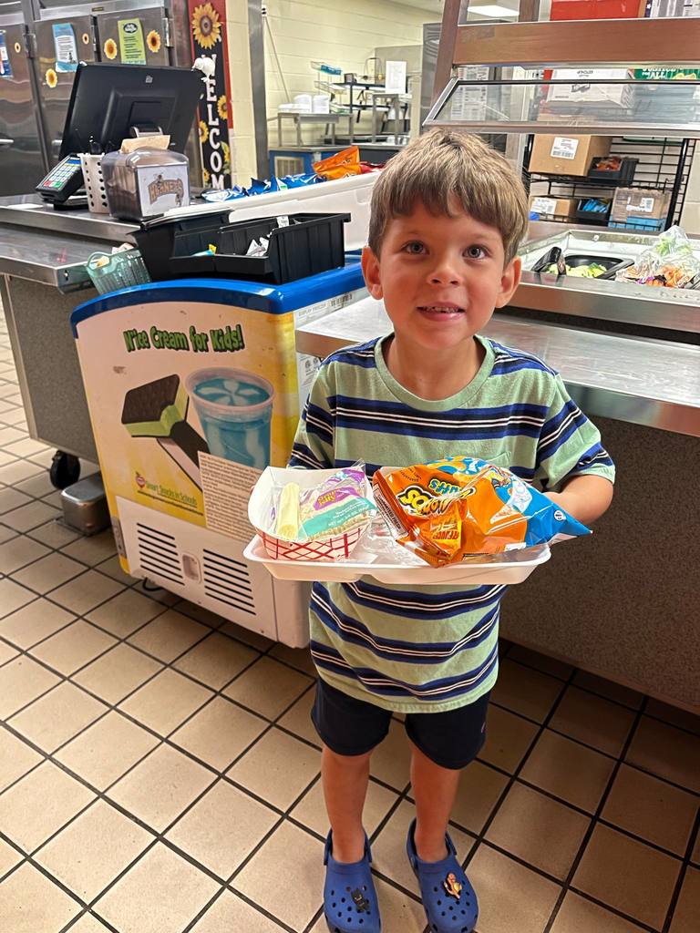 Young boy in a cafeteria holding a tray with packaged food, chips, and milk, smiling for the camera.