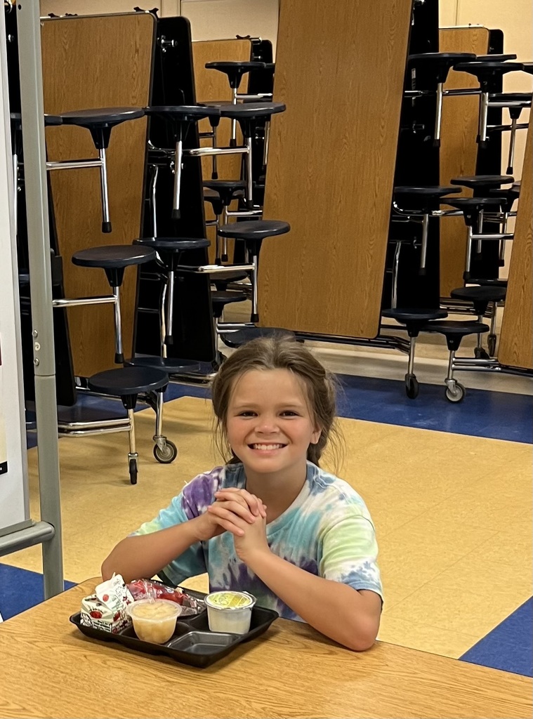 Young girl sitting at a school cafeteria table, smiling, with a tray of fruit, yogurt, and milk in front of her.