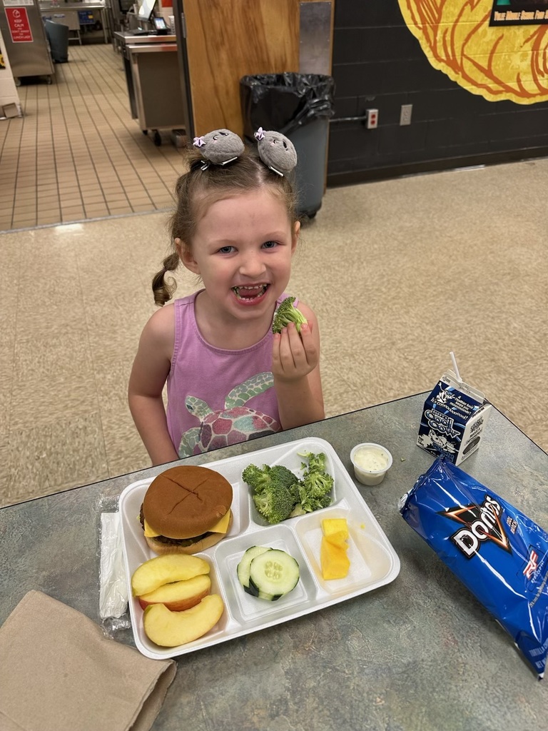 Young girl in a cafeteria smiling while eating broccoli, with a lunch tray containing a cheeseburger, fresh vegetables, fruit, chips, and milk.