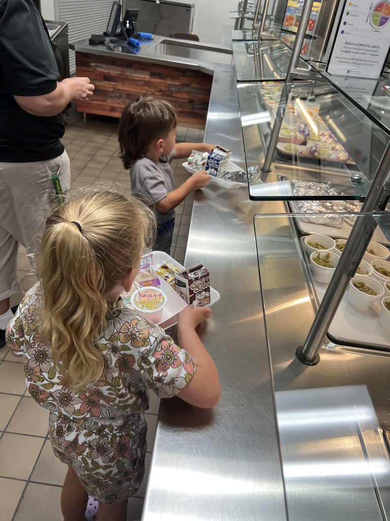 Two young children standing at a school cafeteria serving line selecting milk and food items for lunch.