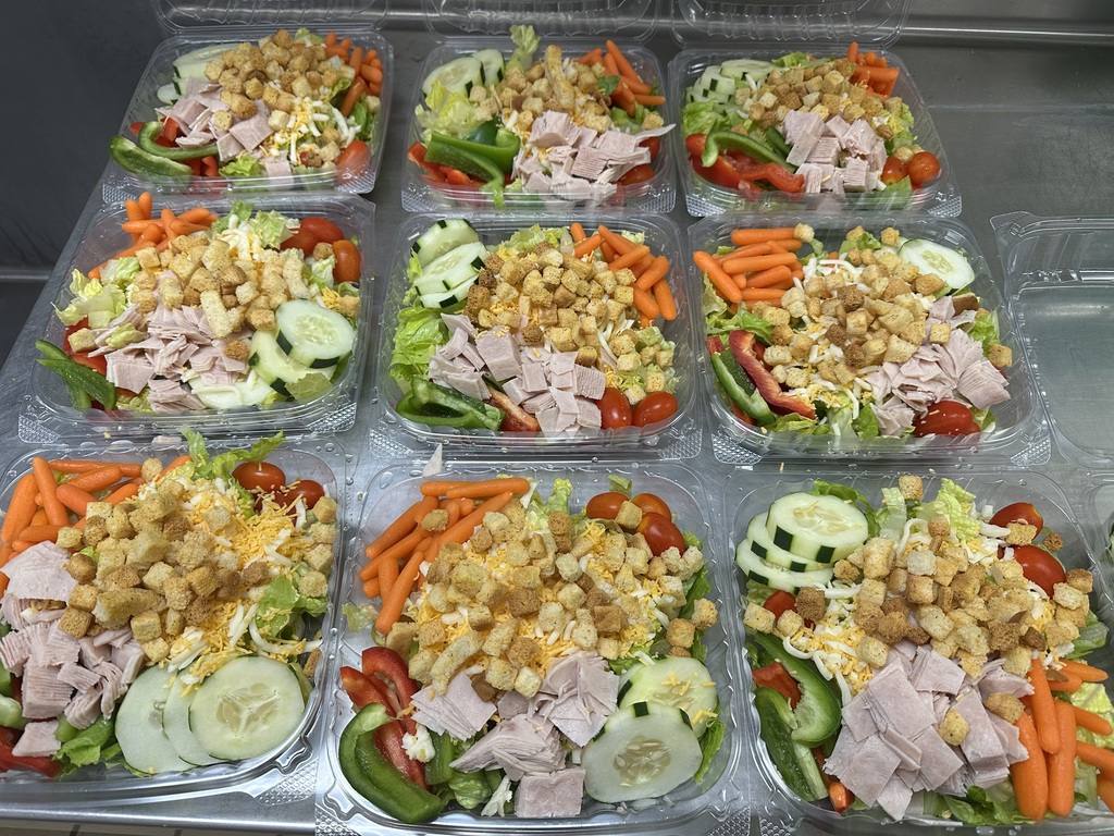 Nine prepared chef salads with turkey, shredded cheese, croutons, tomatoes, cucumbers, carrots, and bell peppers in clear plastic containers.