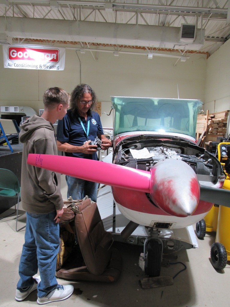 Two people standing in front of a small airplane in a workshop, one holding a mechanical part while the other observes.