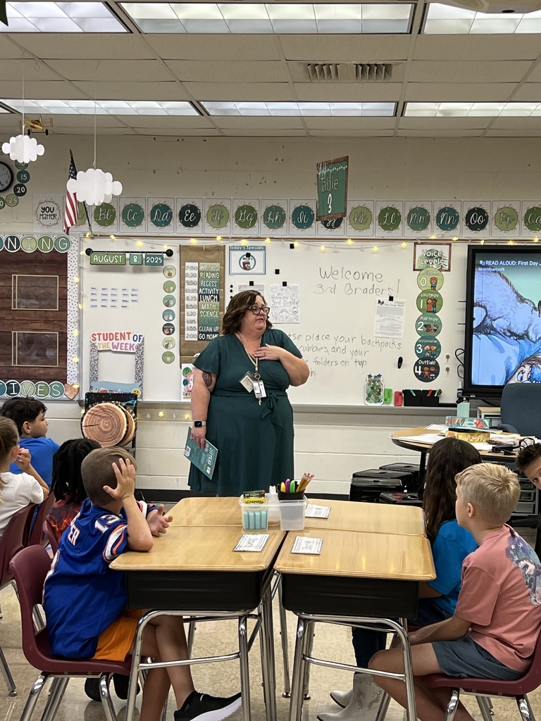 A teacher standing at the front of a third-grade classroom speaking to students seated at desks on the first day of school.
