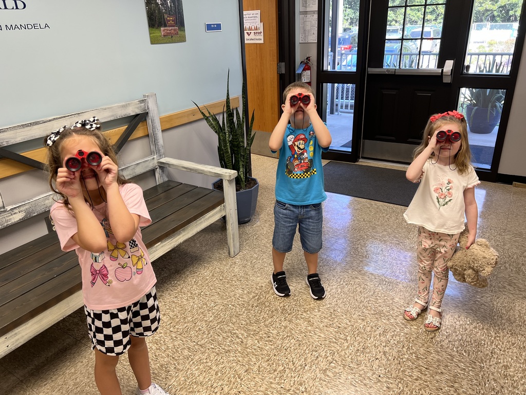 Three young children standing inside a school hallway looking through red toy binoculars, one holding a stuffed animal.