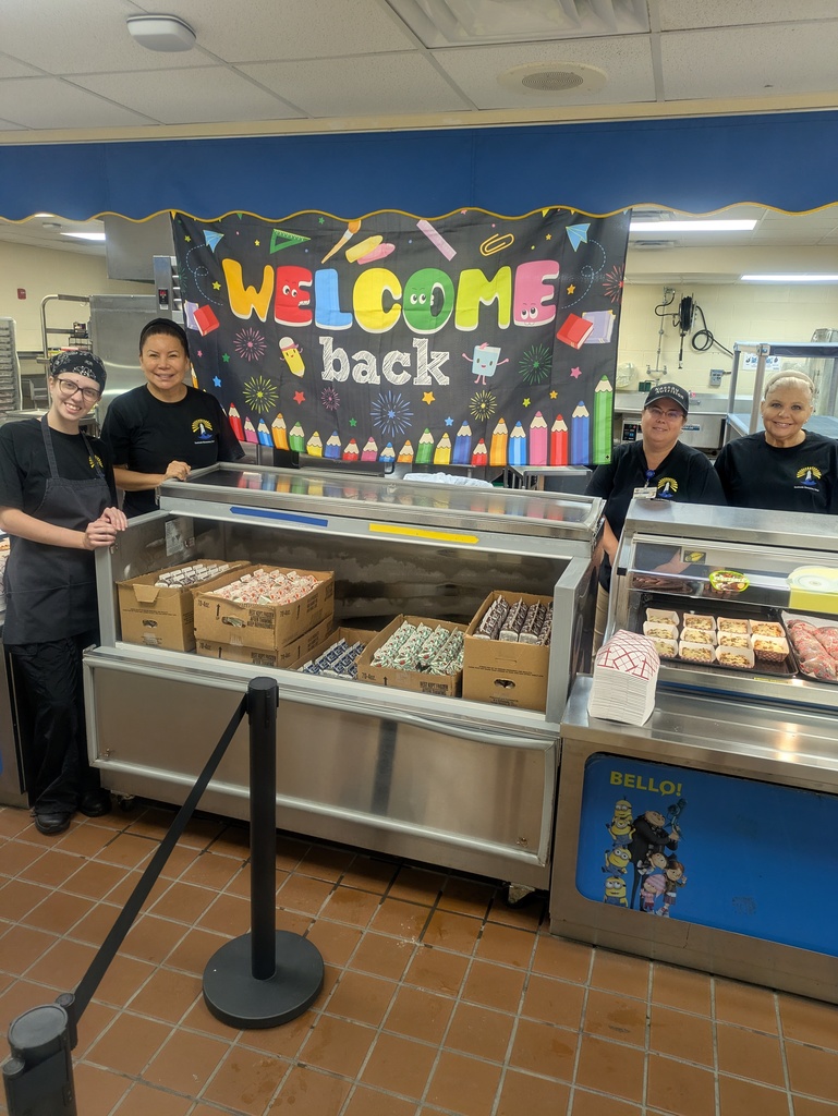 Four cafeteria staff members standing behind a serving counter filled with milk cartons and breakfast items, in front of a colorful "Welcome Back" sign.