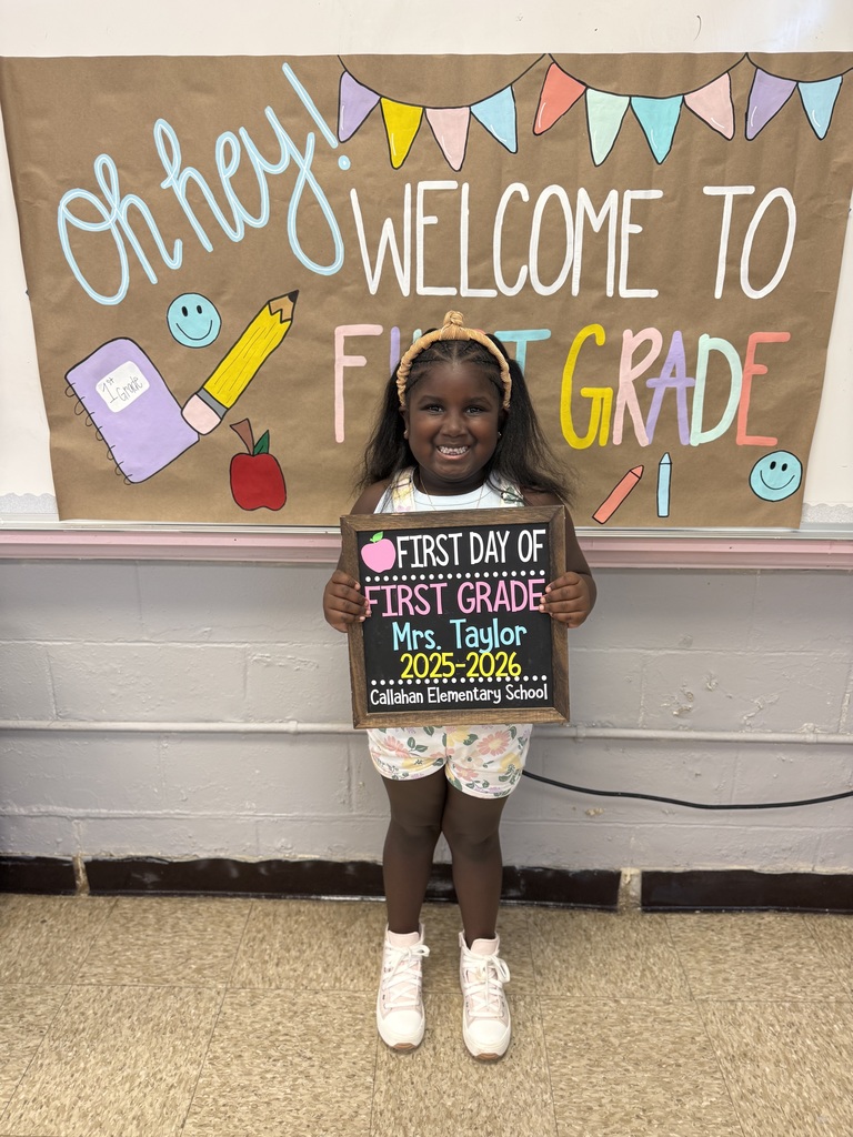 Young girl standing in front of a colorful "Welcome to First Grade" sign, holding a chalkboard sign for her first day of school.