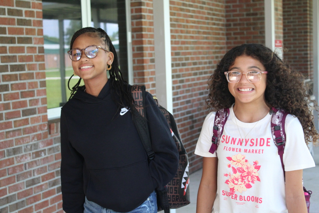 Two middle school students smiling outside near a brick building, wearing backpacks on the first day of school.