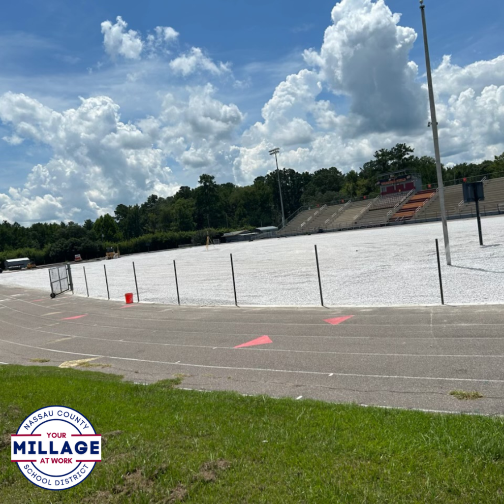 View from the stadium bleachers overlooking the West Nassau High School football field under construction, with a white gravel base in place where the turf will be installed. The school building and scoreboard are visible in the background. Nassau County School District “Your Millage at Work” logo in the bottom left.