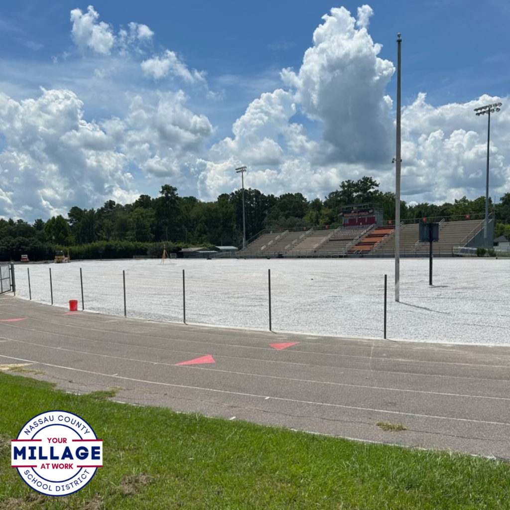 Ground-level view of the West Nassau High School football field under construction, with a white gravel surface and empty stadium seating in the background. Nassau County School District “Your Millage at Work” logo in the bottom left.