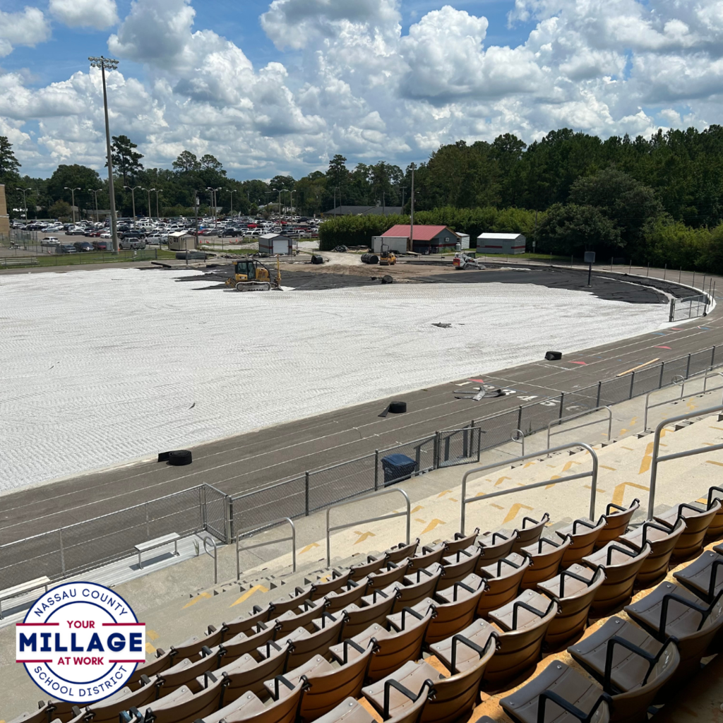 View from the stadium bleachers overlooking the West Nassau High School football field under construction, with a white gravel base in place where the turf will be installed. The school building and scoreboard are visible in the background. Nassau County School District “Your Millage at Work” logo in the bottom left.