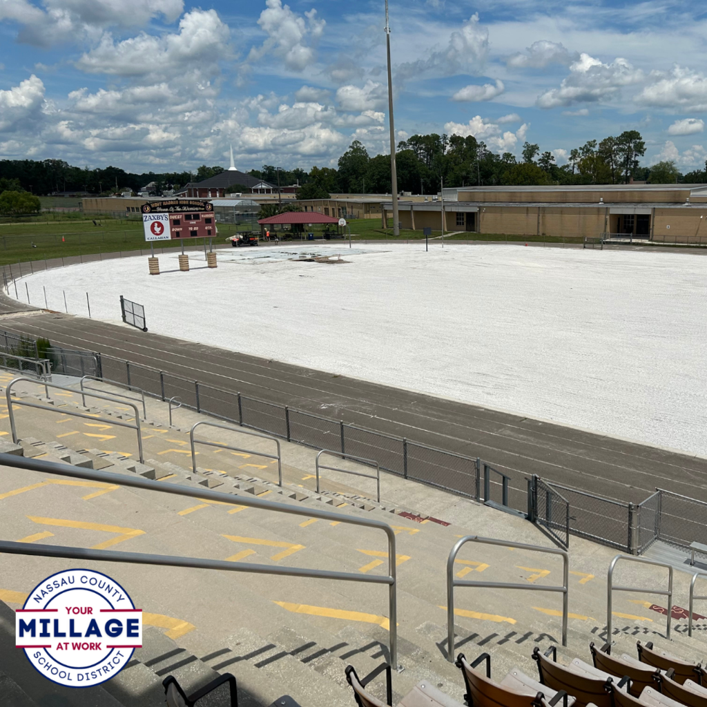 View from the stadium bleachers overlooking the West Nassau High School football field under construction, with a white gravel base in place where the turf will be installed. The school building and scoreboard are visible in the background. Nassau County School District “Your Millage at Work” logo in the bottom left.