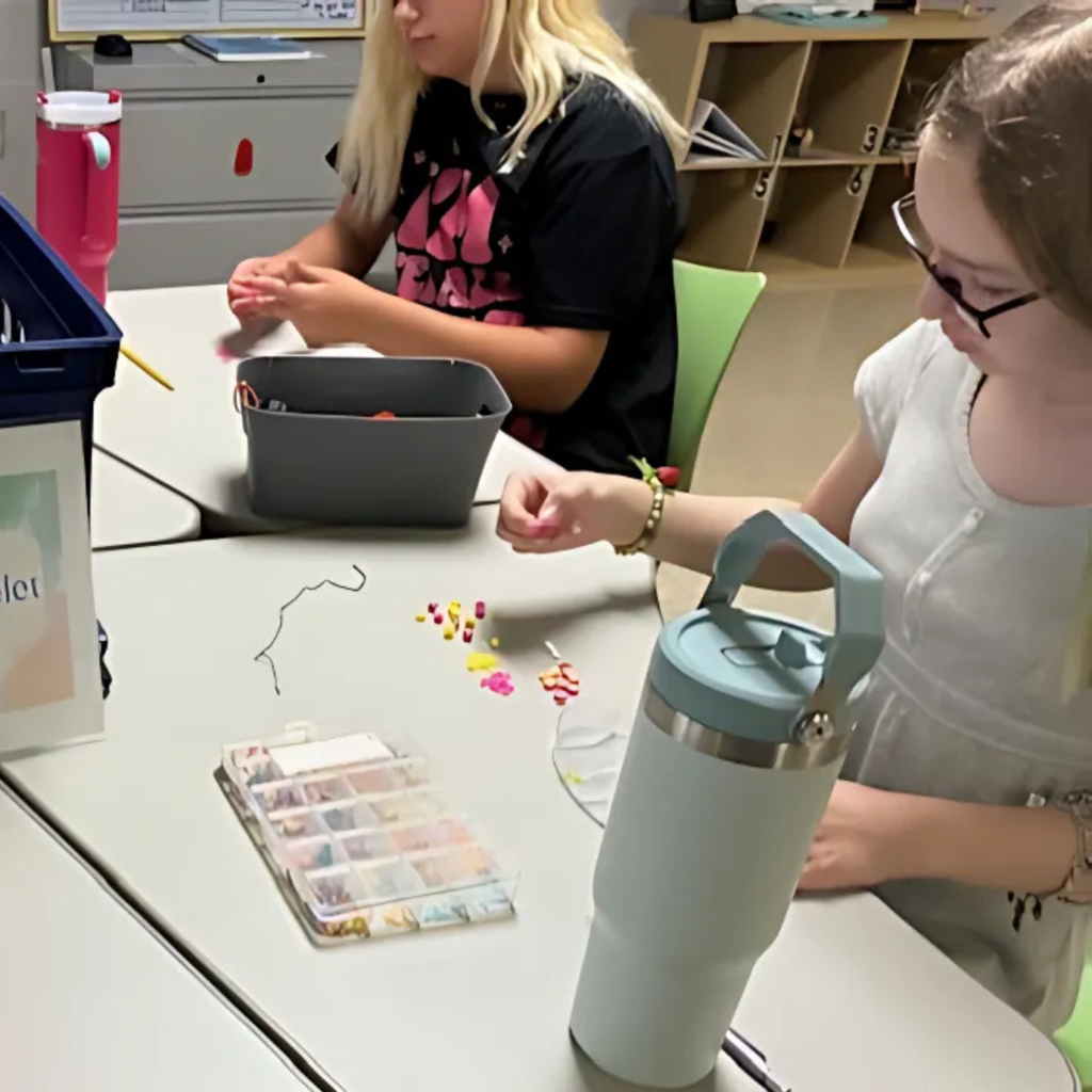 Two students sitting at a desk beading bracelets with string and organizing supplies from a bead kit.