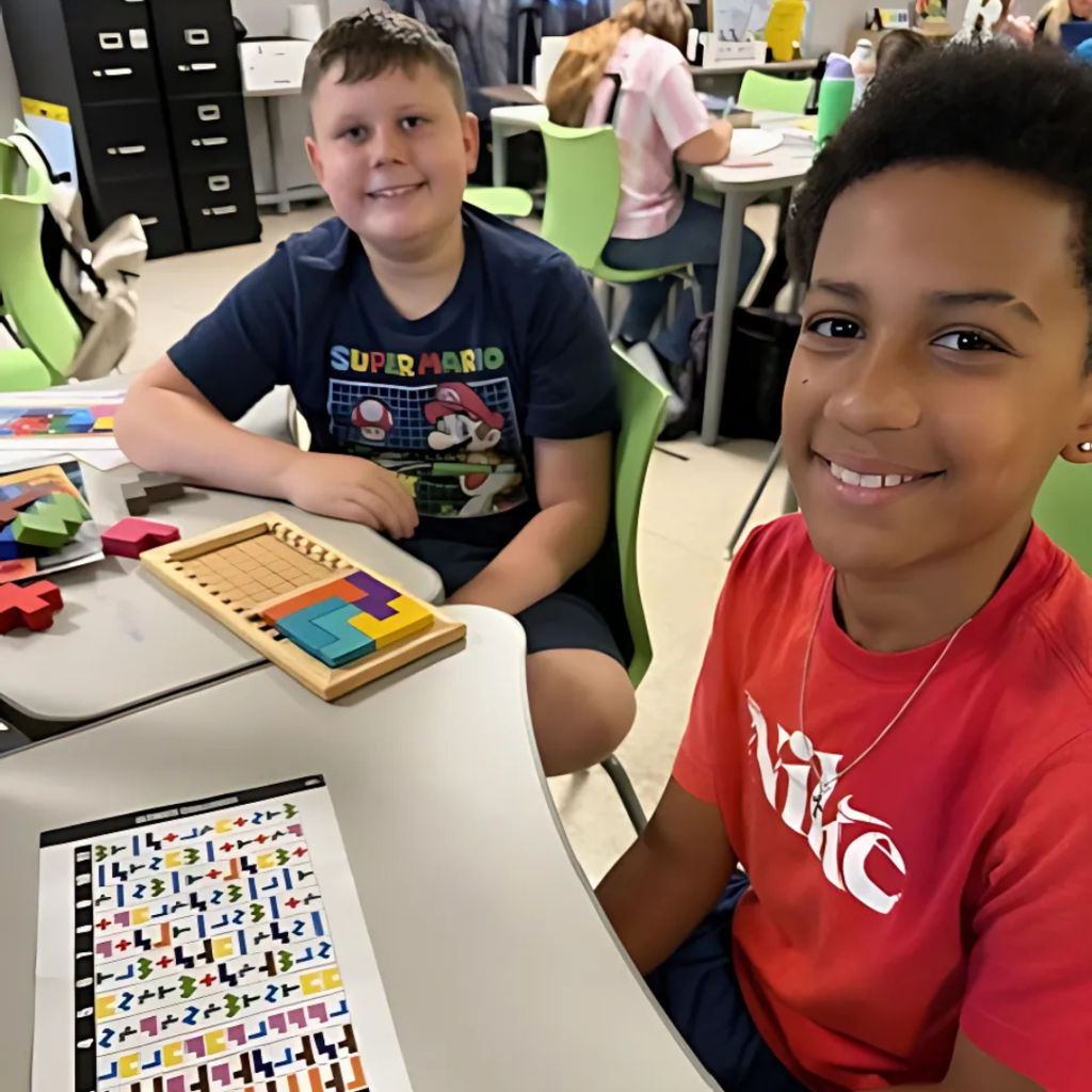Two boys smiling at the camera while working on a wooden puzzle activity at their desks.