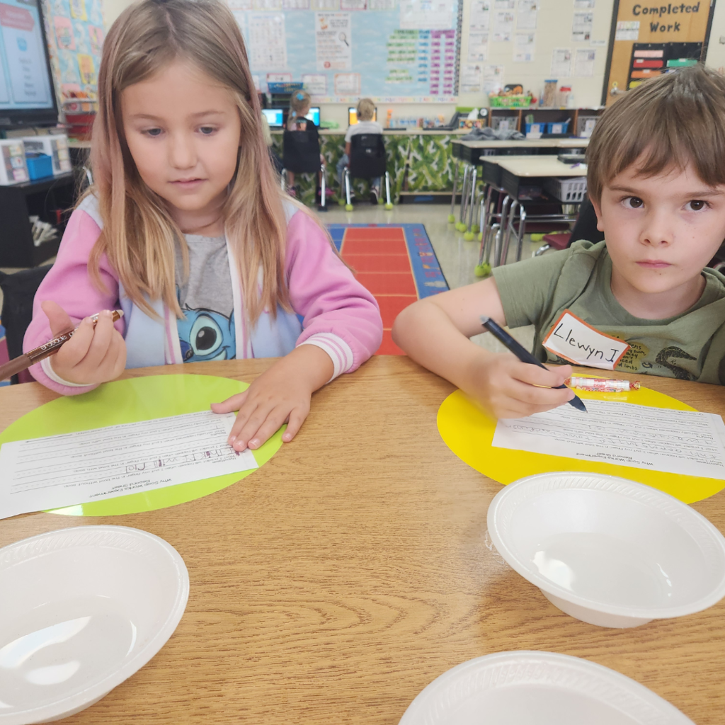 Two students write observations on a worksheet as part of a science lesson at their classroom table with plastic bowls nearby.
