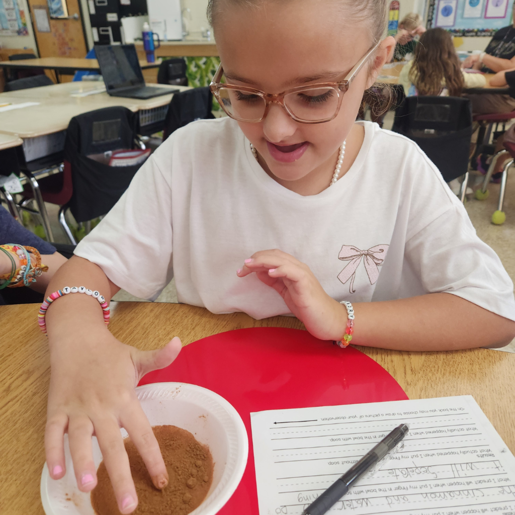 A student smiles while pressing her finger into a bowl filled with cinnamon and water during a soap science experiment.