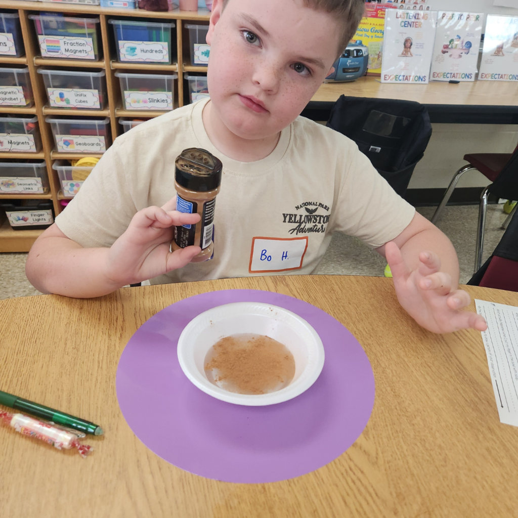 A student named Bo poses with a container of cinnamon next to a bowl of water and spice mixture during a hands-on experiment.