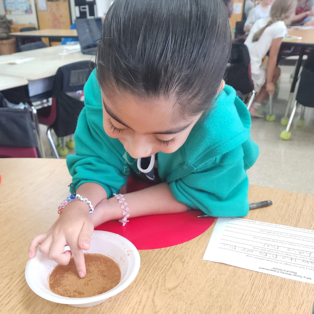A student in a green sweatshirt leans over a bowl, touching the surface of a cinnamon-water mixture during a soap experiment.