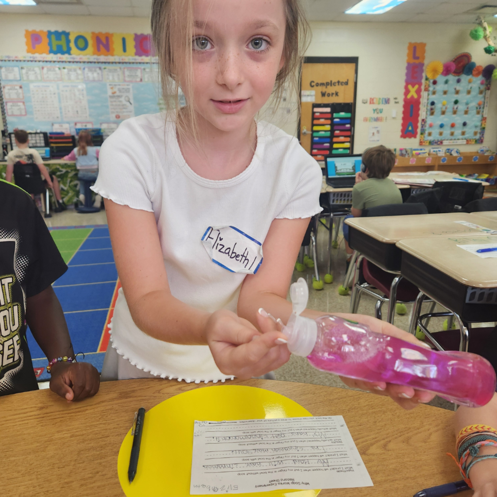 A student named Elizabeth holds a bottle of pink liquid soap while participating in a classroom science experiment.