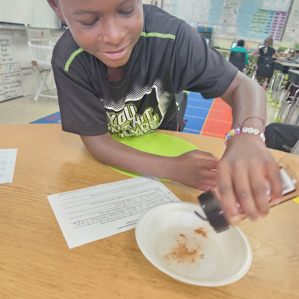A young student smiles while sprinkling a powdered substance into a bowl of water during a classroom science experiment.