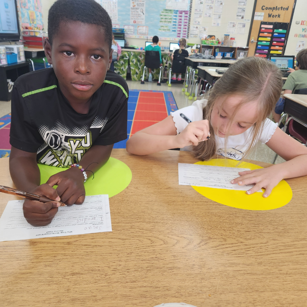 Two students sit at a desk working on a science experiment worksheet together in a bright, welcoming classroom.