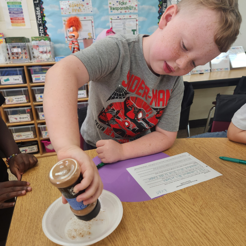 A student in a Spider-Man shirt pours powder into a bowl while participating in a science activity about soap in a colorful classroom.