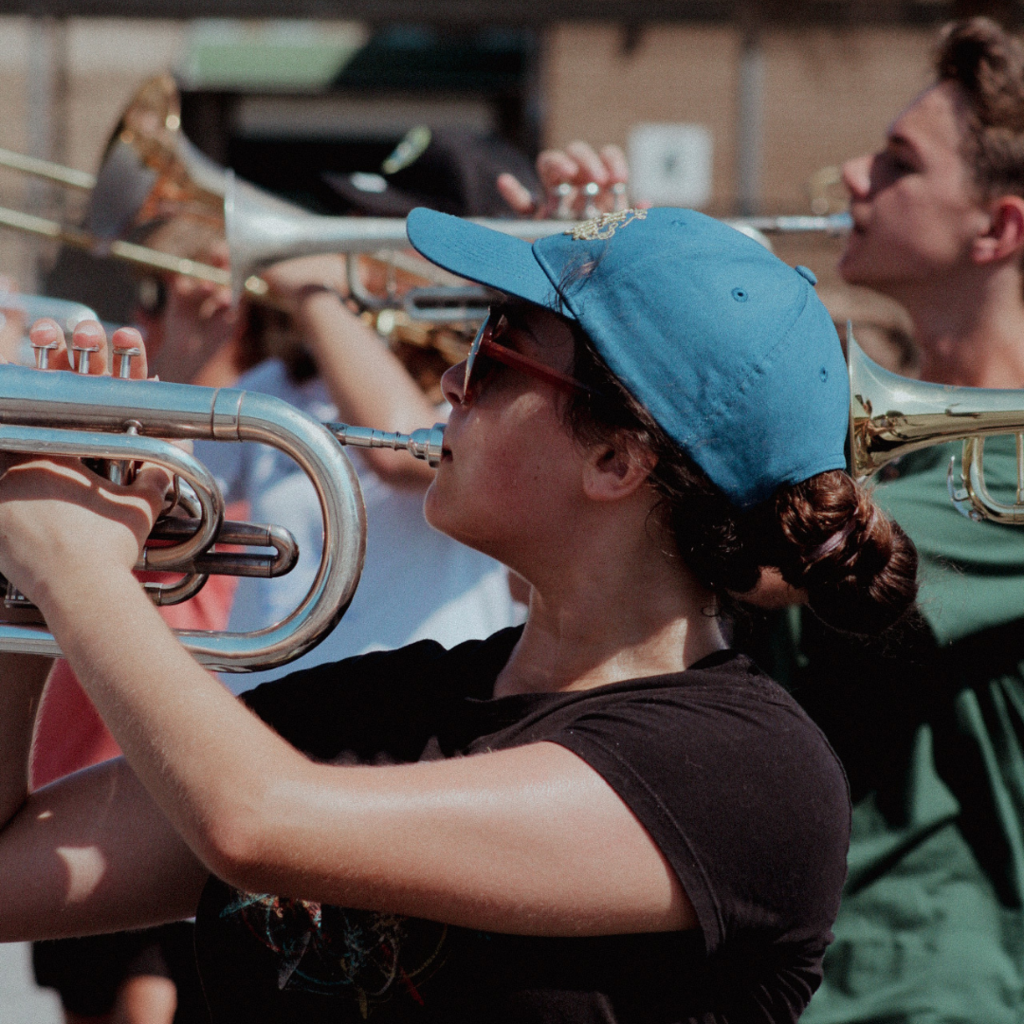 A student wearing sunglasses and a blue cap plays trumpet during a sunny outdoor band practice, surrounded by other brass players.