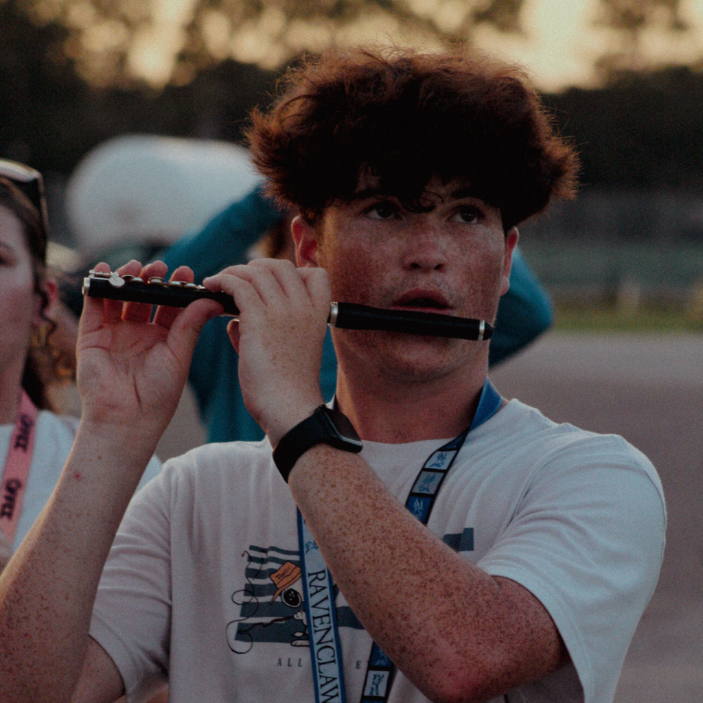 A student focuses while playing the piccolo during evening band practice, wearing a Ravenclaw shirt and a lanyard.