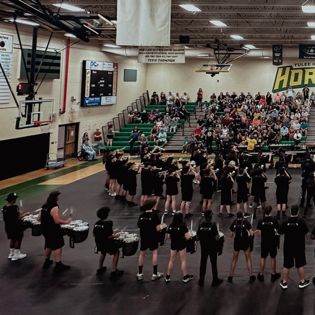 The Yulee High School marching band performs in the school gym as a large crowd of families and supporters watch from the bleachers.