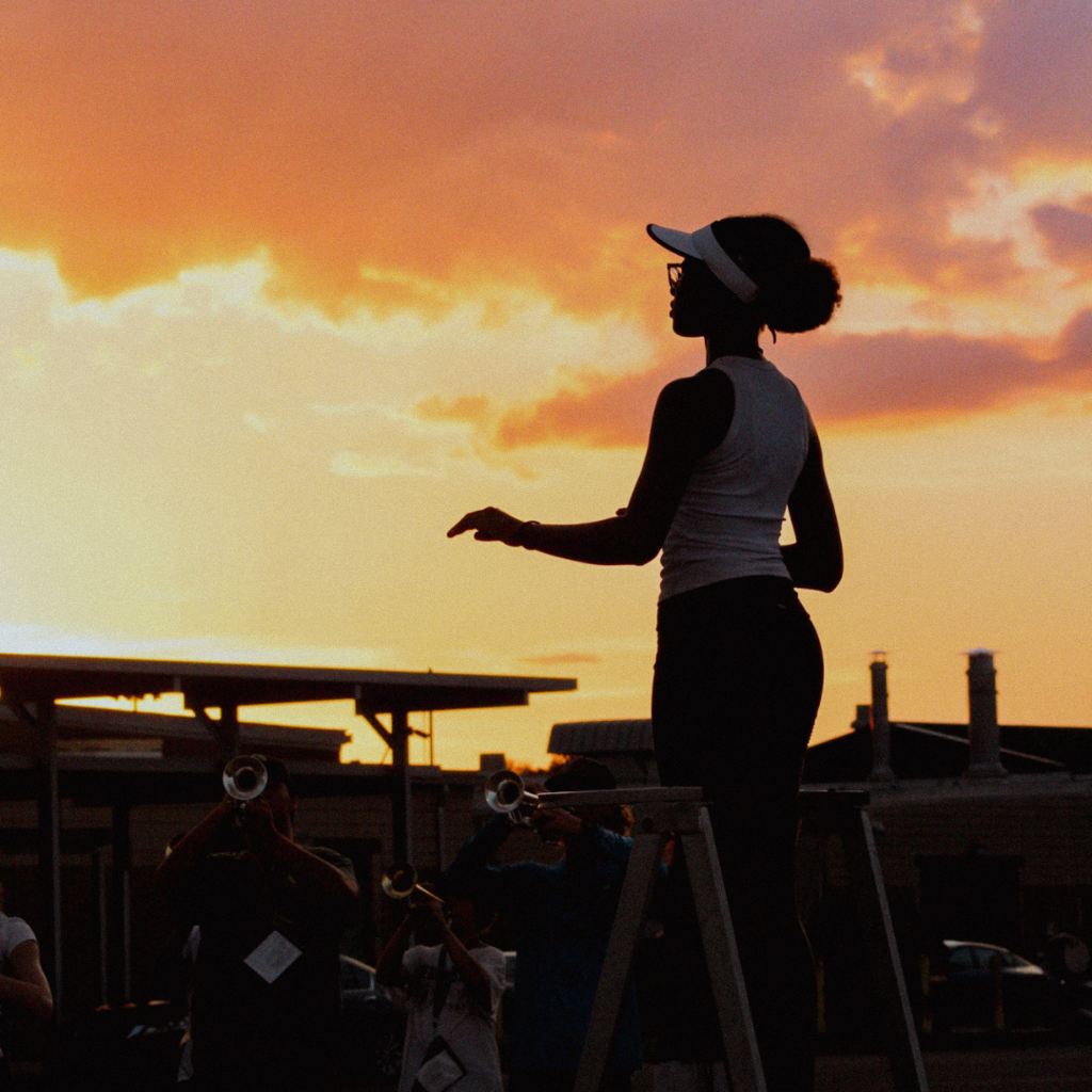 A student drum major stands on a podium silhouetted against a vivid sunset sky, leading band members during an outdoor rehearsal.