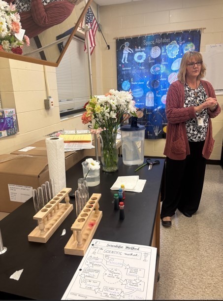 A teacher standing beside a table filled with flowers, test tubes, and science supplies, preparing for the Flower Lab activity. A large poster about the space alphabet hangs on the wall behind.