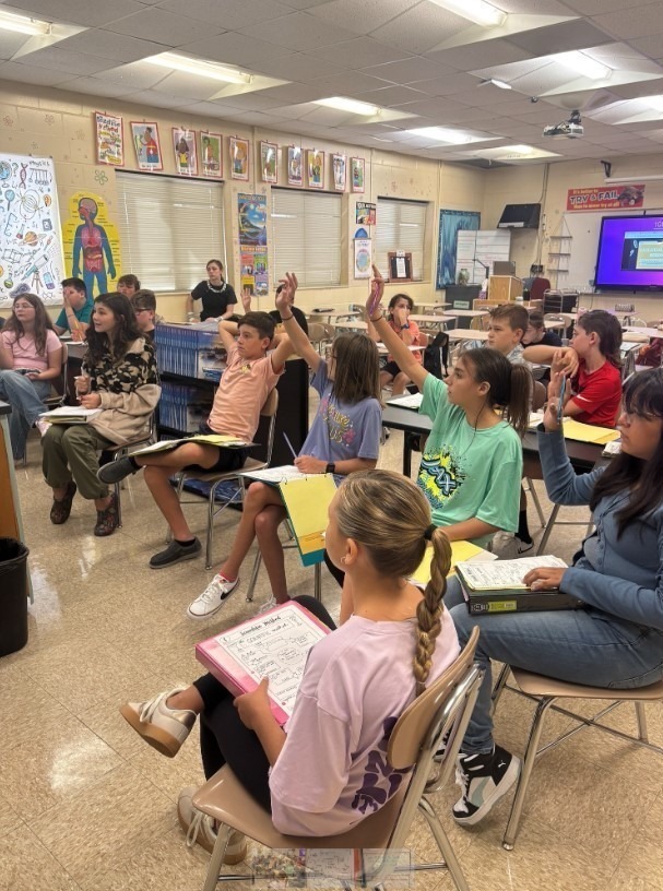 A classroom of sixth grade students seated at desks, many raising their hands to answer a question during a science lesson. Notebooks and binders with the Scientific Method are visible on desks.