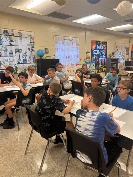 A group of sixth grade students seated at long tables with notebooks and pencils, listening attentively during a science lesson. The walls are decorated with posters and a science-themed curtain.