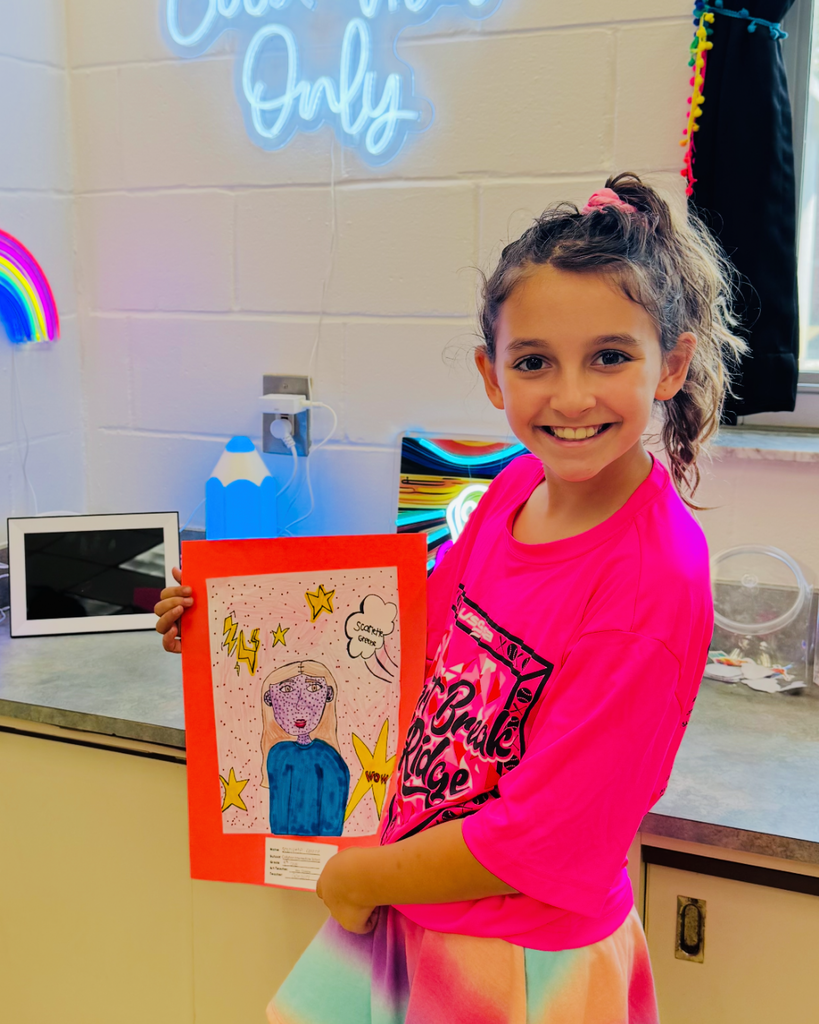 A student holds a self-portrait in the style of Pop Art, featuring a comic-style drawing with bright colors, speech bubbles, and lightning bolts on a pink background.