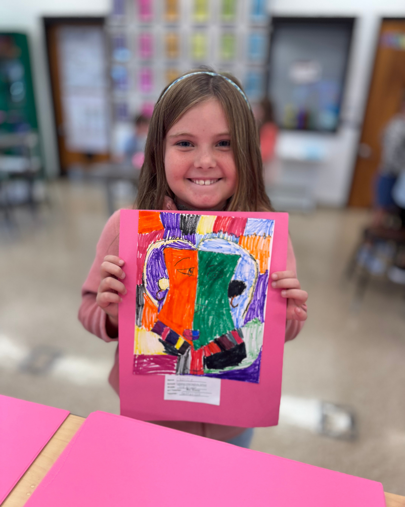 A student holds a Cubism-inspired self-portrait, filled with bold colors and fragmented shapes in orange, green, purple, and black, mounted on pink paper.