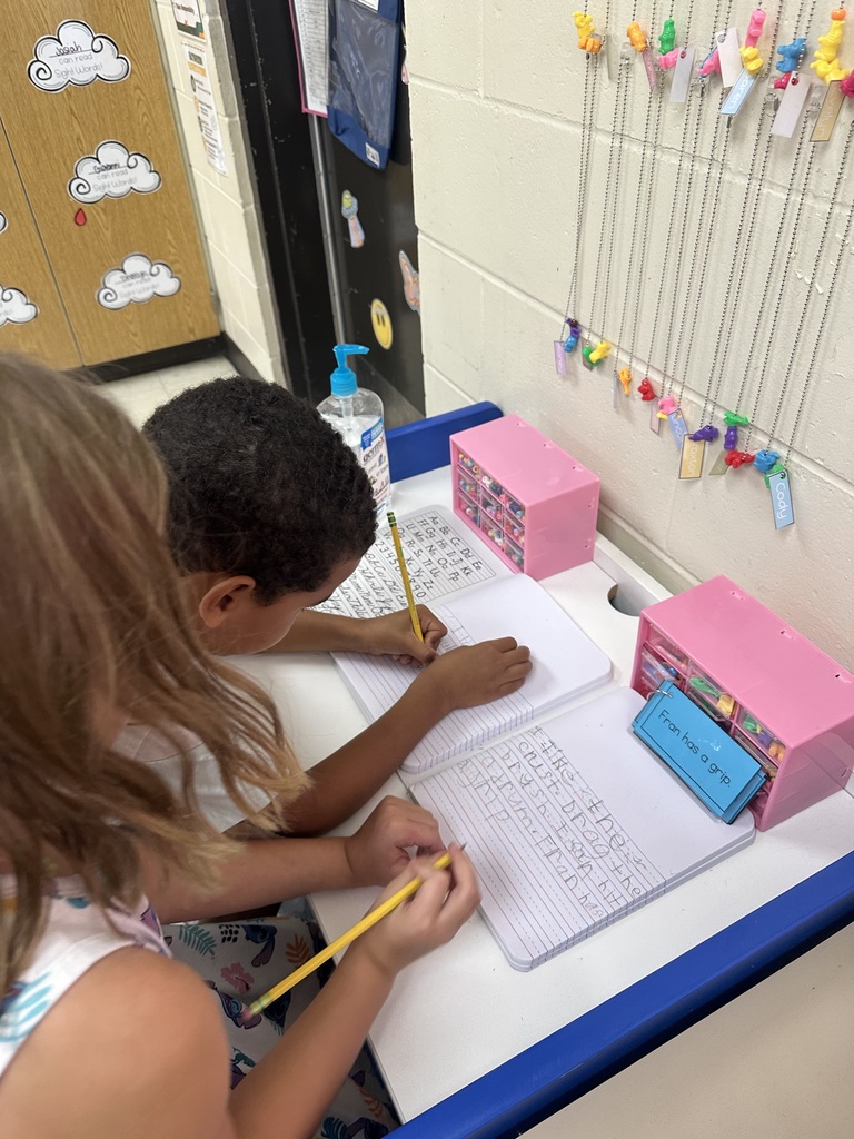 Two students work side by side at a desk, writing in composition notebooks. A set of small drawers and bright keychains with names hang on the wall nearby.