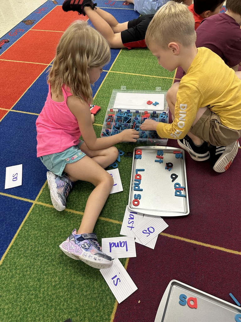 Two first grade students sit on a colorful classroom carpet working with letter tiles and word cards. They are arranging letters on a metal tray to build words like “land” and “so.”