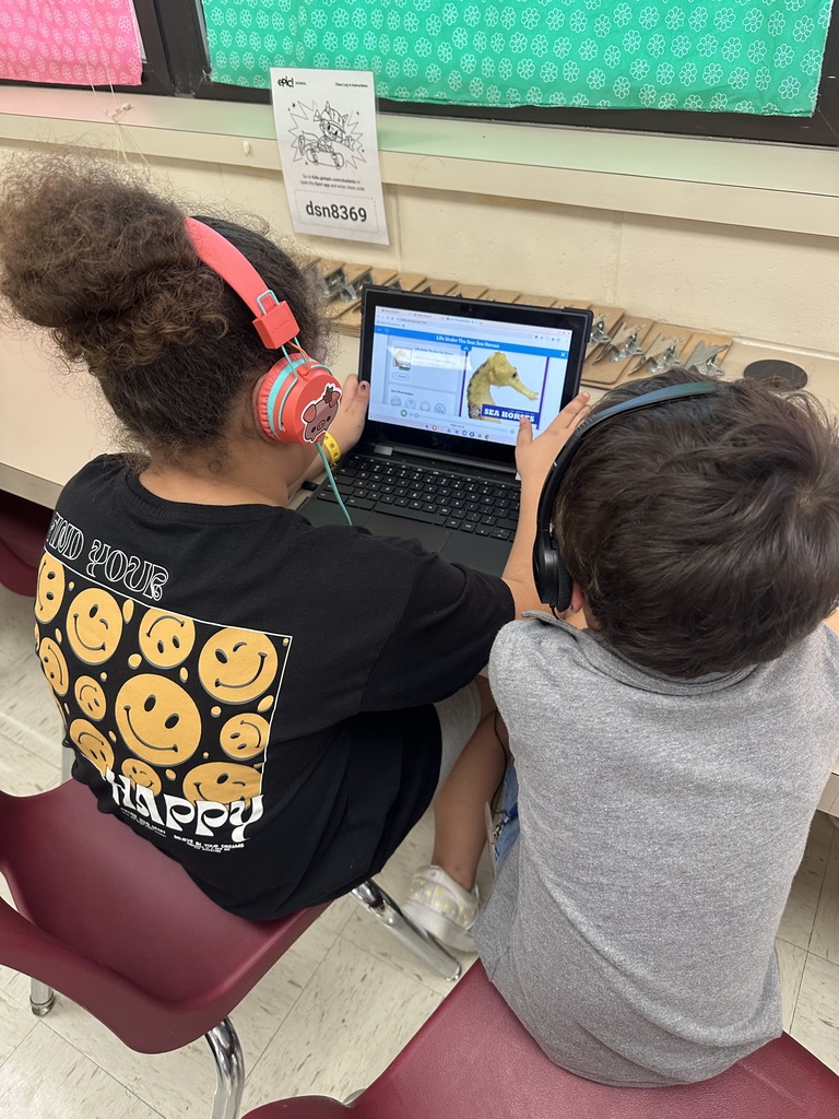 Two students wearing headphones sit at a laptop in the classroom. The screen displays an educational program featuring a seahorse.
