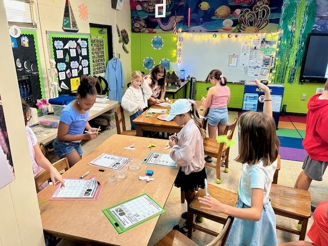 A student stands next to a classroom table and releases a paper “spin-a-copter” into the air while another student observes and records notes.