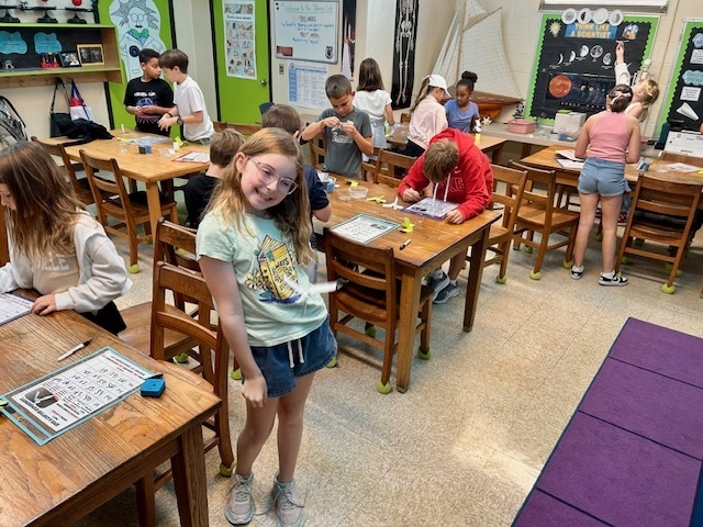 A student smiles at the camera while holding a paper “spin-a-copter.” Behind her, classmates continue building and testing their projects at tables.
