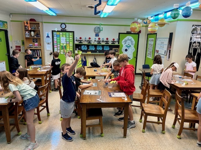 Students in a science classroom work in groups at wooden tables. They are folding and testing paper “spin-a-copters” as part of an experiment.