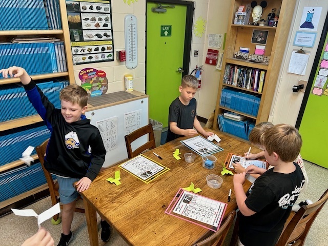 Four students gather around a classroom table. One student holds up a paper “spin-a-copter” while others write on worksheets and prepare their models.