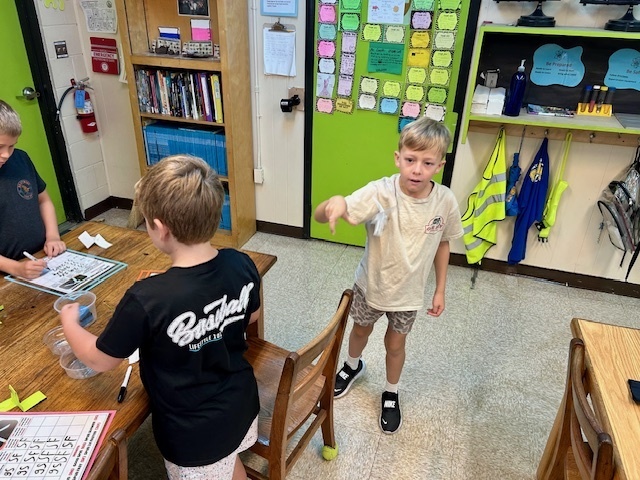 Groups of students work together at tables in a colorful science classroom. Several are folding, testing, and recording results from paper “spin-a-copters.”