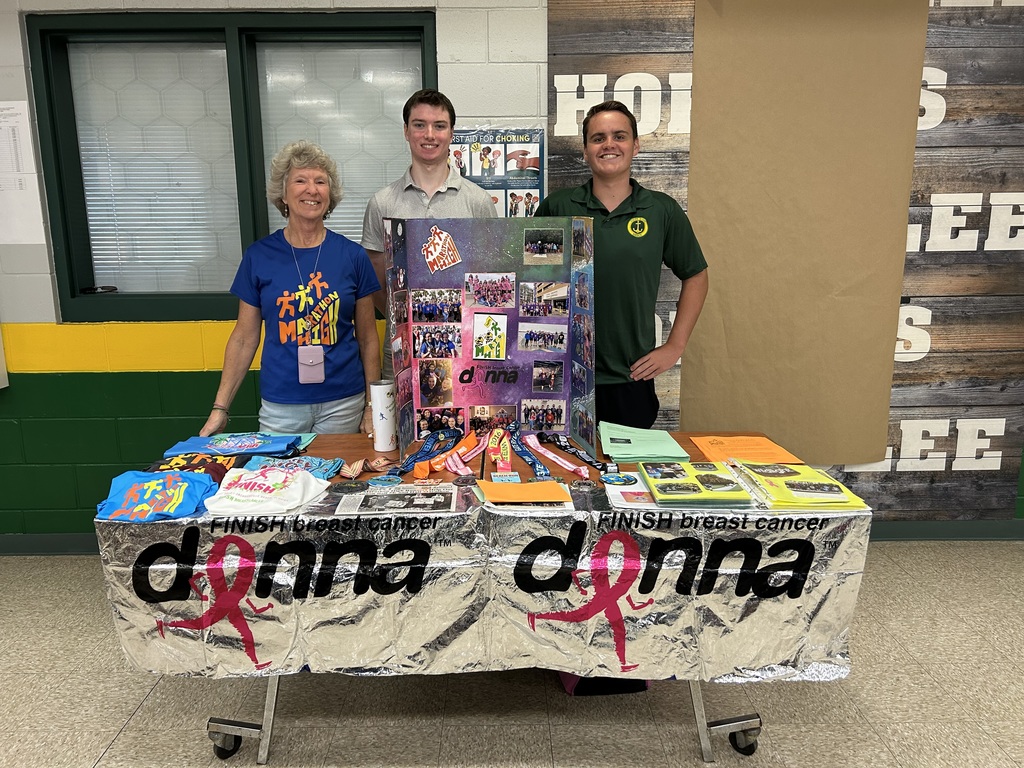 An adult and two students stand behind the Marathon High Running Club display board. The table is filled with medals, race shirts, and flyers promoting training for the Donna Half Marathon.