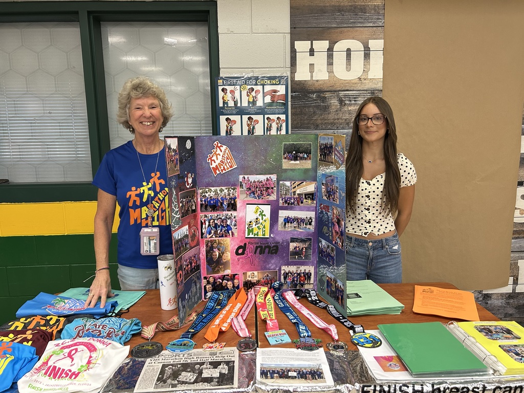 An adult and a student stand behind a colorful display board for Marathon High Running Club. The table is covered with race shirts, medals, flyers, and a banner promoting the Donna Half Marathon to finish breast cancer.