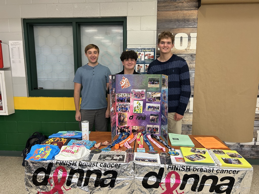 Three students stand behind a Marathon High Running Club display board decorated with photos and medals. The table features t-shirts, flyers, and a silver banner with the Donna Half Marathon logo.