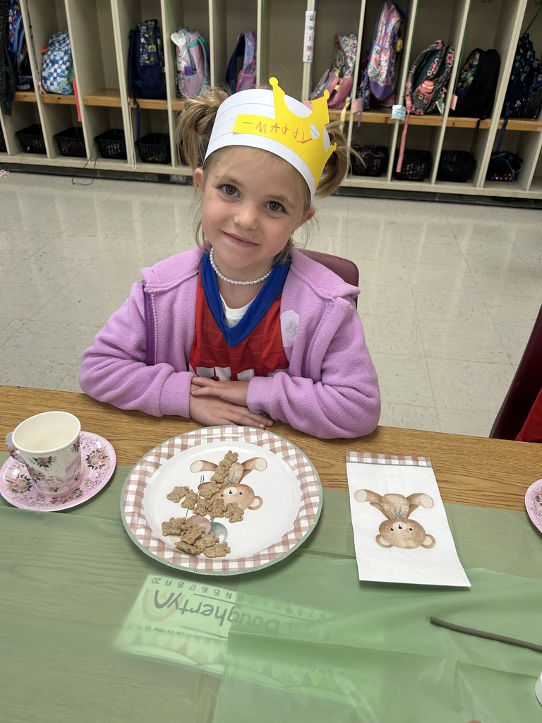 A kindergarten student wearing a yellow crown sits at a decorated table with a plate of teddy bear cookies, a teacup, and a teddy bear napkin.