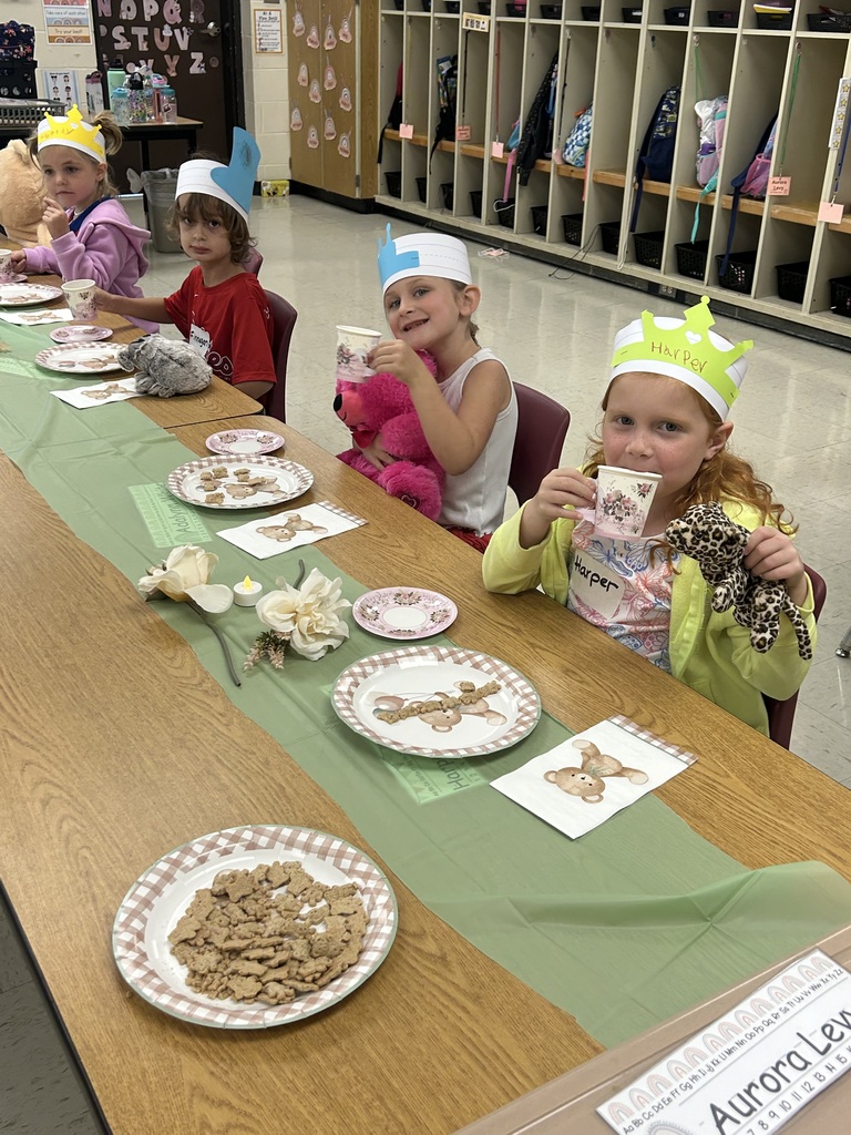 Four kindergarten students wearing paper crowns sit at a table with teddy bear plates and stuffed animals while smiling and drinking from teacups.