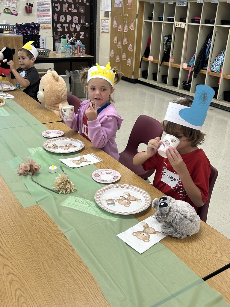 Two kindergarten students wearing paper crowns sip from teacups at a table decorated with flowers, teddy bear plates, and stuffed animals.