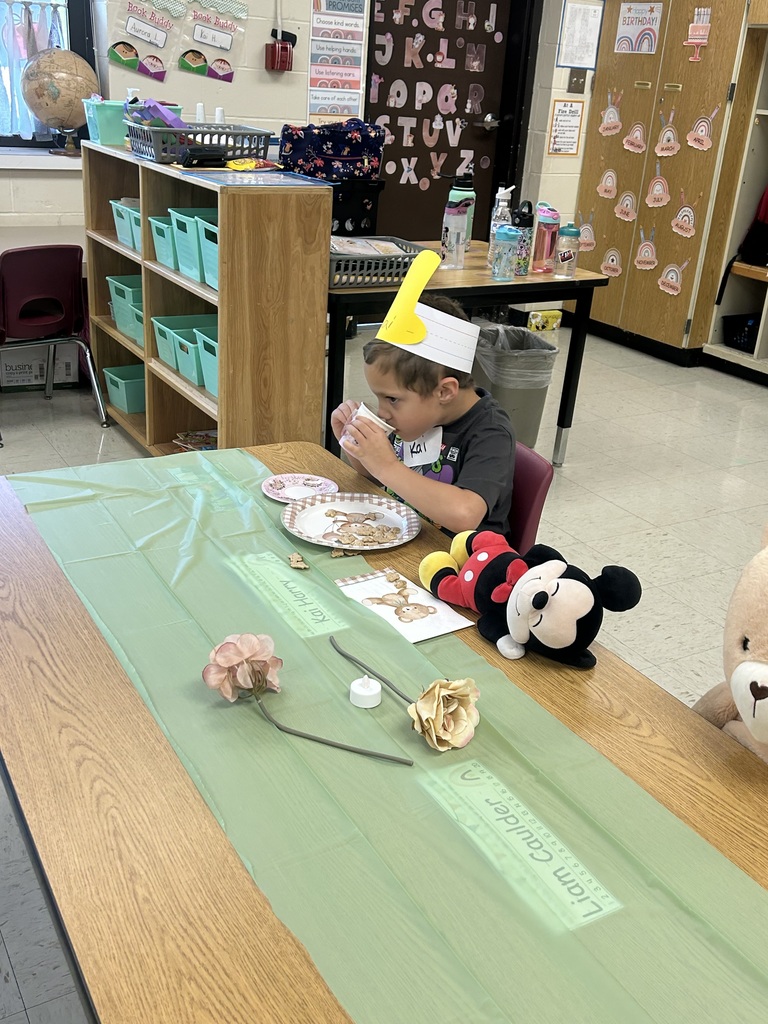 A kindergarten student wearing a yellow paper crown drinks from a teacup at a decorated table with teddy bear plates and stuffed animals.